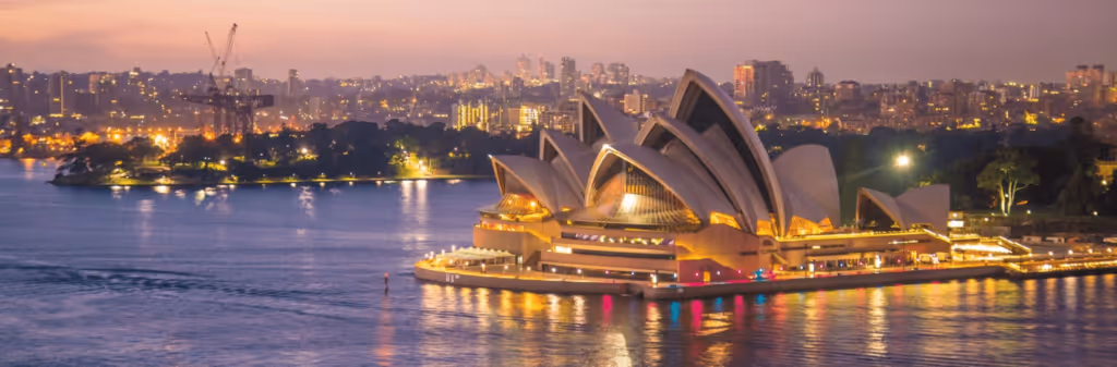 A panoramic view of the Sydney Opera House at dusk, its iconic sails lit up and reflected in the harbour. The illuminated city skyline stands out in the distance, completing the scene.