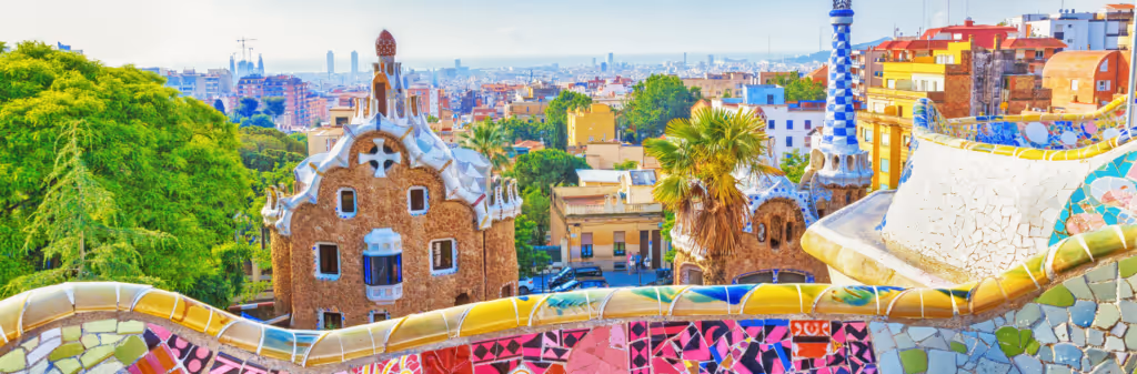 A view from Park Güell in Barcelona. The foreground shows a colorful, mosaic bench with two of the park's iconic buildings. The city and sea are visible in the distance under a clear sky.