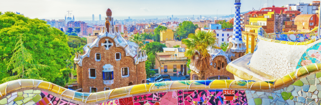 A view from Park Güell in Barcelona. The foreground shows a colorful, mosaic bench with two of the park's iconic buildings. The city and sea are visible in the distance under a clear sky.