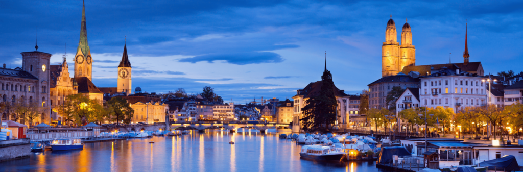 A panoramic view of Zurich at dusk, with the Limmat River and its illuminated old city buildings in the foreground. The Grossmünster church towers and other historic spires are lit up against the twilight sky.