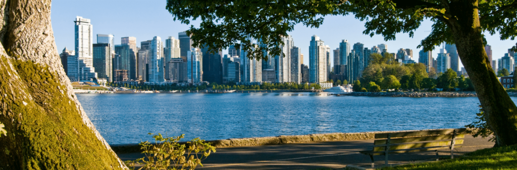 A view of the Vancouver skyline from across the water. Large trees and a park bench frame the foreground, with the city's modern buildings and a green waterfront visible in the background.