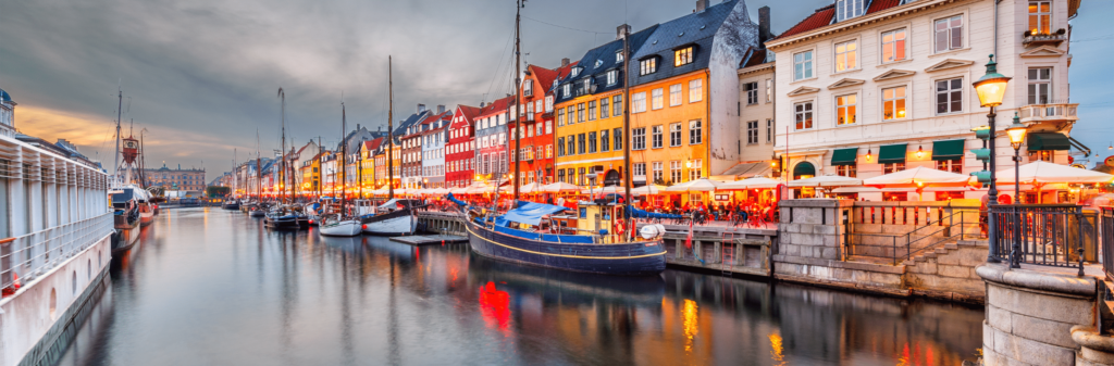 A view of the colorful waterfront buildings along the Nyhavn canal in Copenhagen at dusk. Boats are docked in the harbor, and lights from the buildings and streetlamps reflect in the water.
