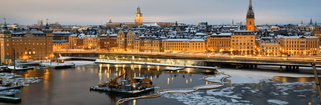 A winter view of Stockholm at dusk. The city's historic buildings are lit up, contrasting with the snowy rooftops and the partially frozen river in the foreground.