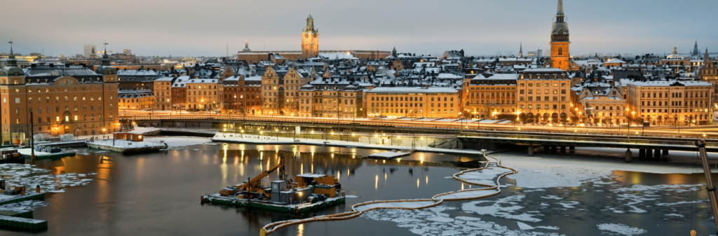 A winter view of Stockholm at dusk. The city's historic buildings are lit up, contrasting with the snowy rooftops and the partially frozen river in the foreground.