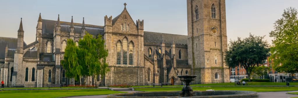 A historic stone cathedral with a tall clock tower and a fountain in a green park.