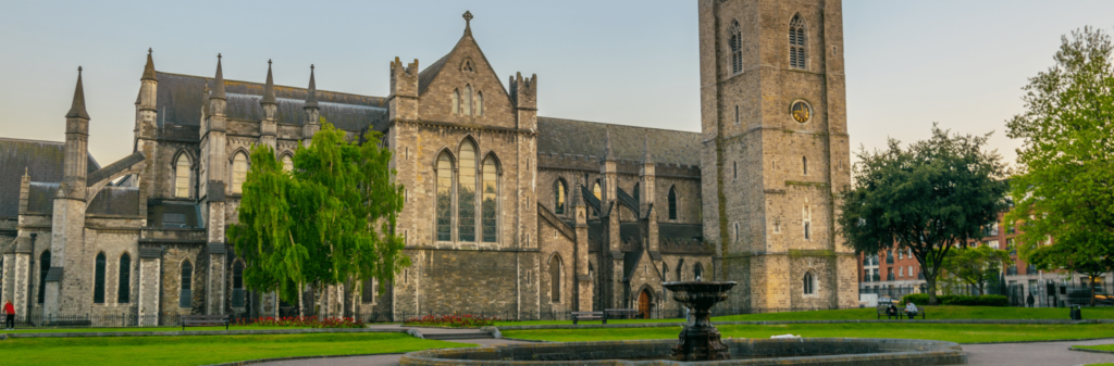 A historic stone cathedral with a tall clock tower and a fountain in a green park.