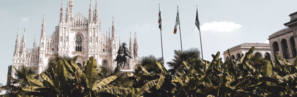 View of Milan Cathedral, a statue, and palm trees with three flagpoles in the foreground.