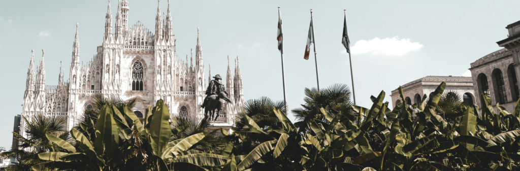 View of Milan Cathedral, a statue, and palm trees with three flagpoles in the foreground.