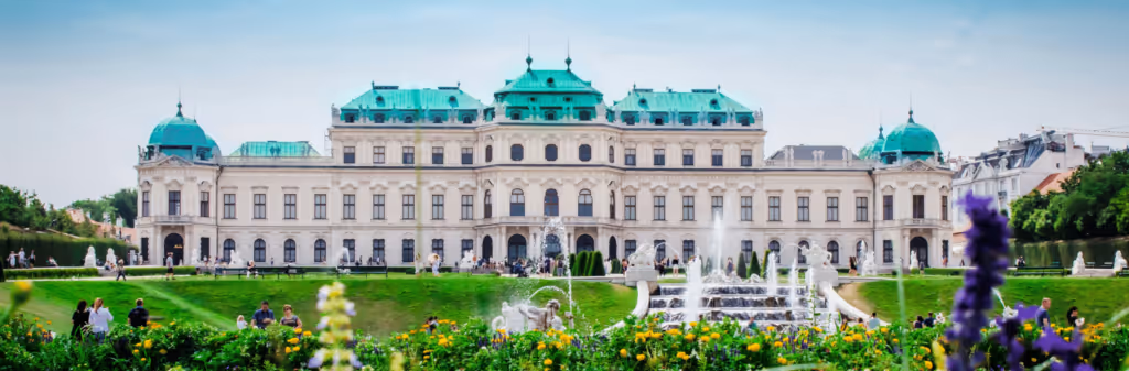 A grand palace with green roofs, gardens, and fountains, crowded with visitors on a sunny day.