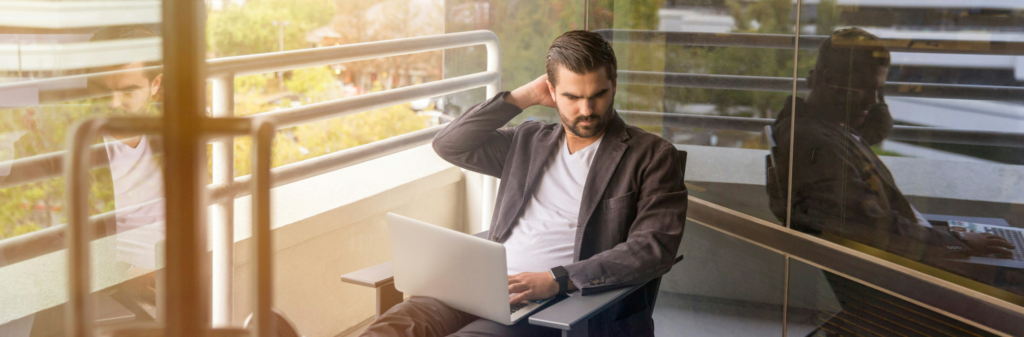 Man sitting on a balcony using a laptop, looking thoughtful with one hand behind his head.