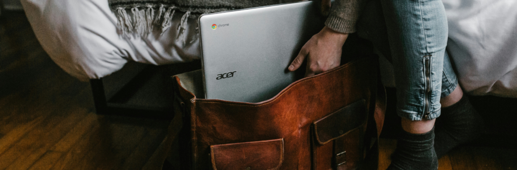 Person placing a silver Acer Chromebook into a brown leather bag beside a bed.