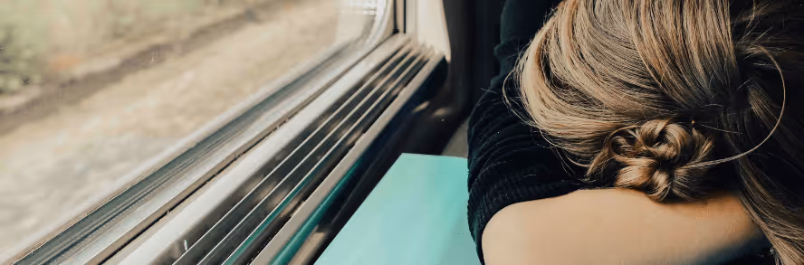 A tired person resting their head on folded arms beside a window, symbolizing exhaustion and burnout often experienced during frequent business travel.