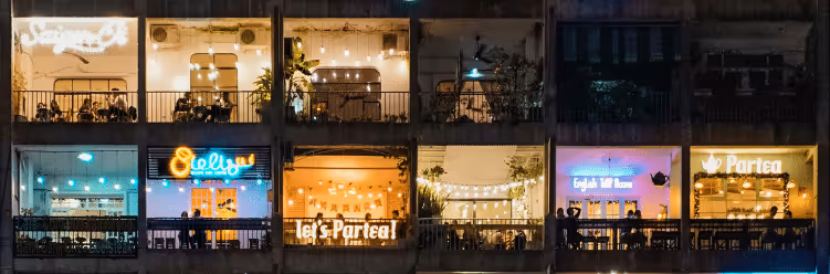 A row of brightly lit cafes and restaurants seen through large windows at night.