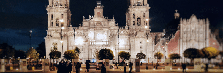 Mexico City Metropolitan Cathedral illuminated at night with people in the foreground and a dark blue sky above.