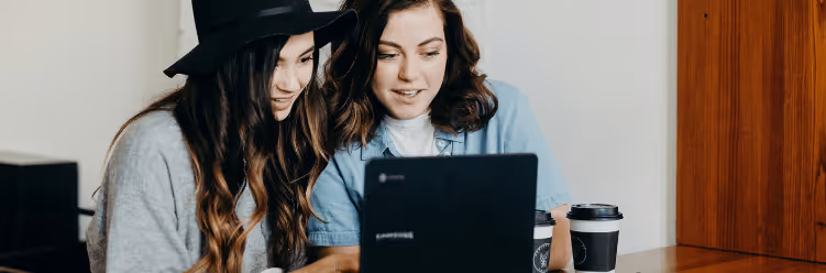 Two female colleagues are seated together in front of a laptop, engaged in conversation about a work task.