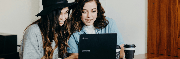 Two female colleagues are seated together in front of a laptop, engaged in conversation about a work task.