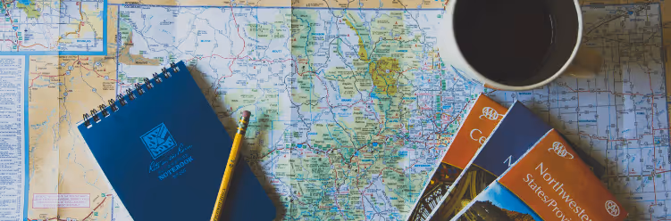 A map with a notebook, pencil, travel guides, and a cup of coffee placed on top.