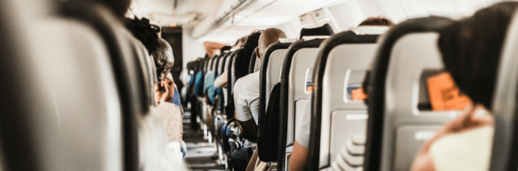 Passengers seated in rows inside an airplane cabin, viewed from behind, with overhead storage compartments visible.