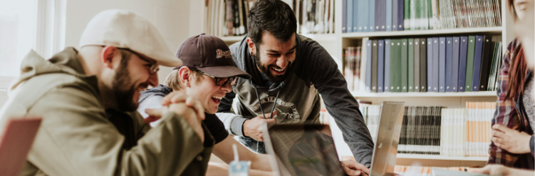 Four people smiling and laughing together while looking at laptops in a library or office setting.