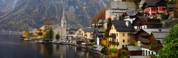 A lakeside village with colorful houses and a tall church tower is nestled against a mountain backdrop under cloudy skies, offering an idyllic setting for sustainable business travel with its serene environment and eco-friendly accommodations. | Dyme