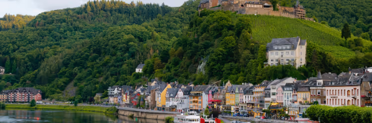 A riverside view of a town with colorful buildings nestled below a forested hill topped with a historic fortress, where the river and tour boat encourage sustainable business travel in the foreground. | Dyme