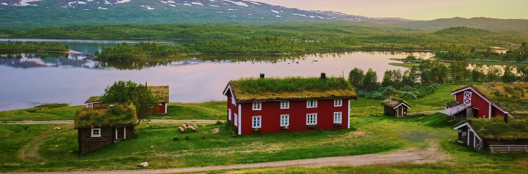 Red houses with grass roofs beside a calm lake and surrounded by greenery, with mountains in the background, offer an idyllic escape for sustainable business travel enthusiasts seeking serenity and eco-friendly accommodations. | Dyme