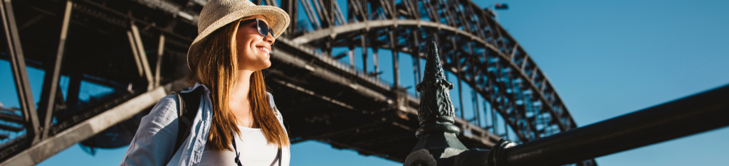 Woman in sunglasses and hat smiling in front of a large steel bridge on a sunny day.