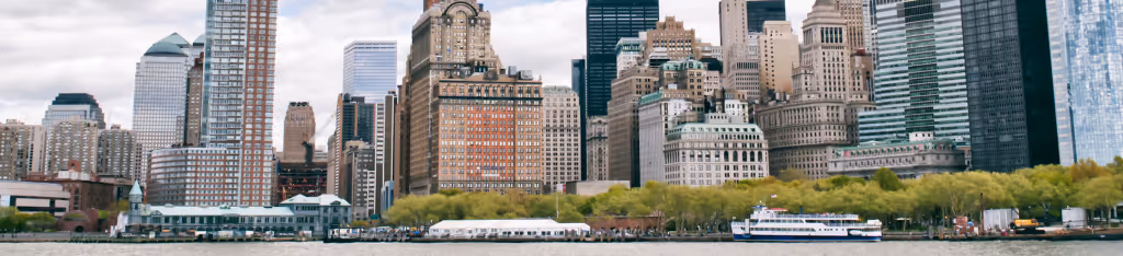 Skyscrapers and waterfront buildings in a city skyline, with trees and a boat on the river in front.
