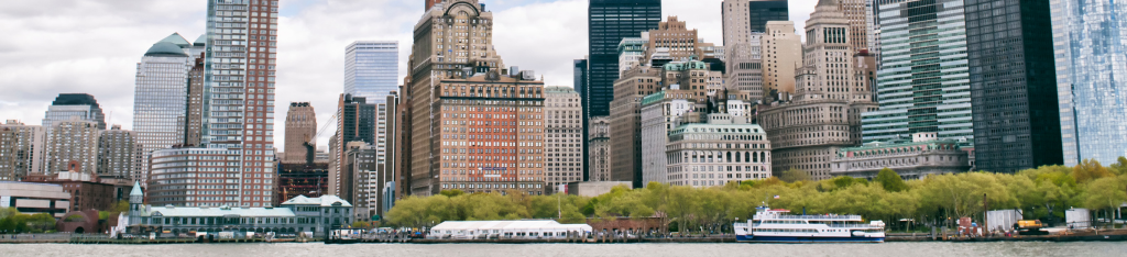 Skyscrapers and waterfront buildings in a city skyline, with trees and a boat on the river in front.