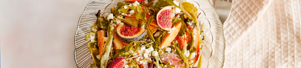 A glass plate of salad with figs, greens, carrots, and crumbled cheese on a light tablecloth.