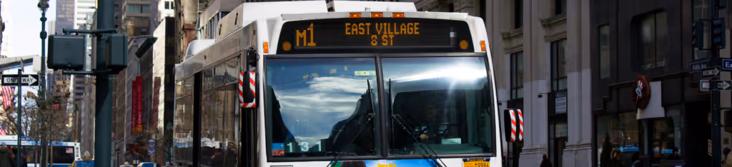 A city bus displaying M1 East Village 8 St on its sign drives down a busy urban street.