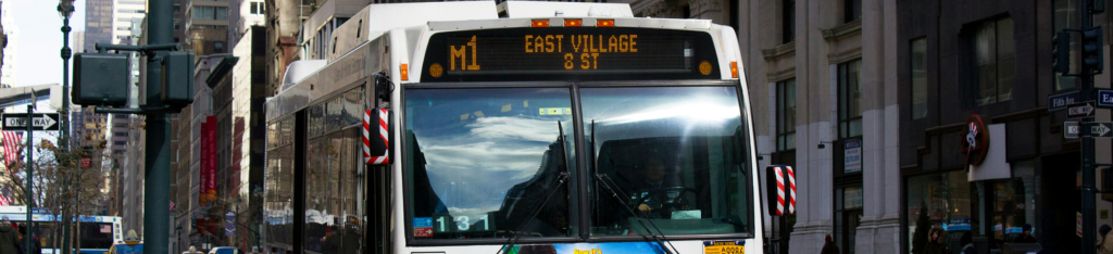 A city bus displaying M1 East Village 8 St on its sign drives down a busy urban street.