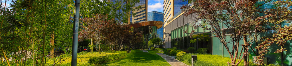 A modern office building with glass walls, surrounded by green trees, shrubs, and a neatly trimmed lawn.