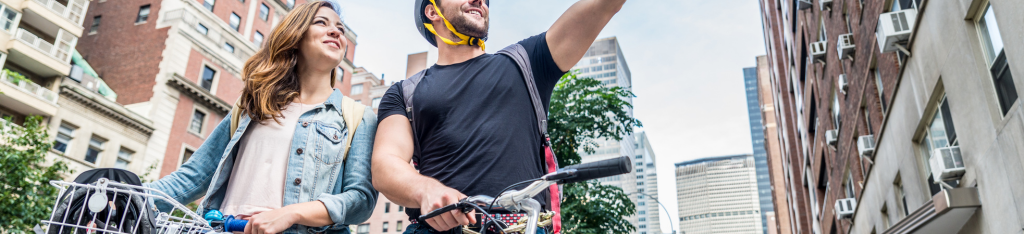 A man and woman with bikes smile and look up on a city street, surrounded by tall buildings.