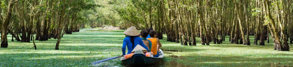 Three people in a boat paddle through a lush, green flooded forest with sunlight filtering through the trees.