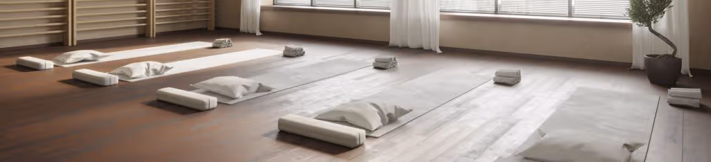 A bright yoga studio with mats, bolsters, and blankets neatly arranged on a wooden floor by large windows.