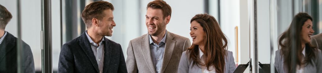 Three business professionals smiling and talking together in an office hallway.