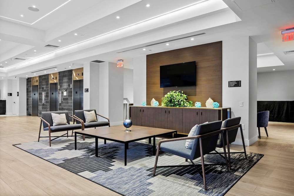 Modern hotel lobby with chairs, tables, a rug, plants, and a TV mounted on the wall above a cabinet.