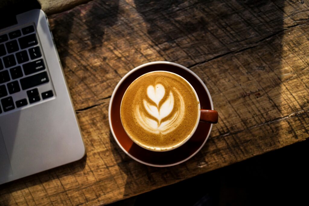 A cup of latte with leaf latte art sits on a rustic wooden table next to a laptop.