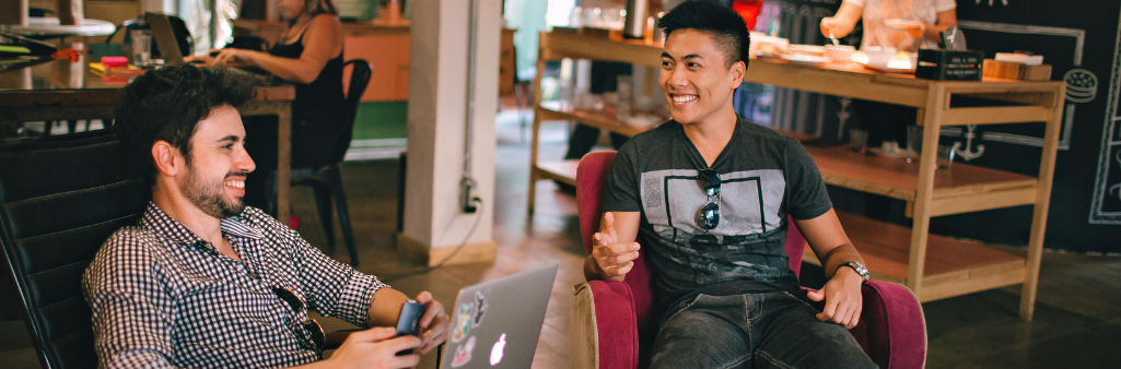 Two men smiling and chatting in a cozy café, one holding a phone, the other sitting with a laptop.
