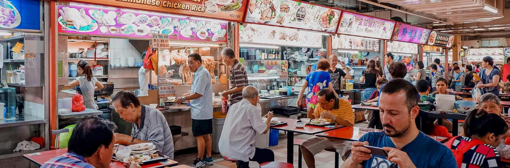 People eating and ordering food at a busy indoor hawker center with various colorful food stalls.