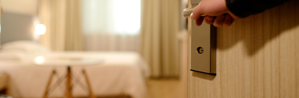 Hand opening a door to a softly lit hotel room with a bed and table visible in the background.