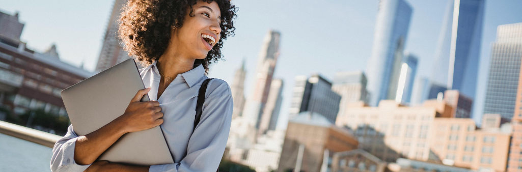 Smiling woman holding a laptop outdoors with a city skyline in the background.