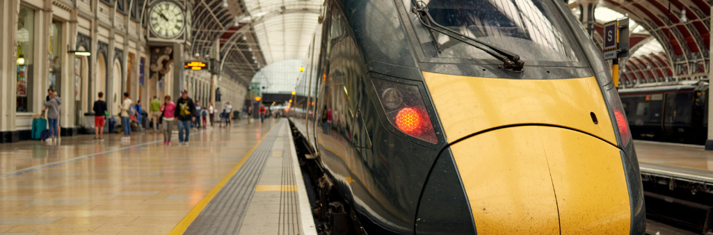 A modern train at a busy indoor station with passengers and a large clock in the background.