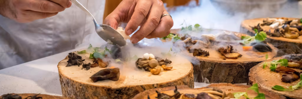 A chef plating gourmet mushrooms and greens on wooden slabs surrounded by dramatic food smoke.