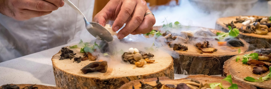 A chef plating gourmet mushrooms and greens on wooden slabs surrounded by dramatic food smoke.