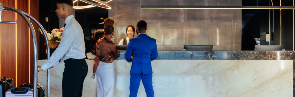 Three people stand at a hotel reception desk while a staff member pushes a luggage cart nearby.