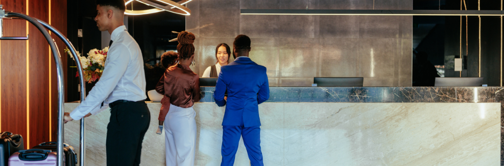 Three people stand at a hotel reception desk while a staff member pushes a luggage cart nearby.