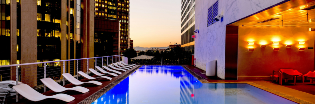 Rooftop pool at sunset, surrounded by lounge chairs and city buildings with lights glowing.