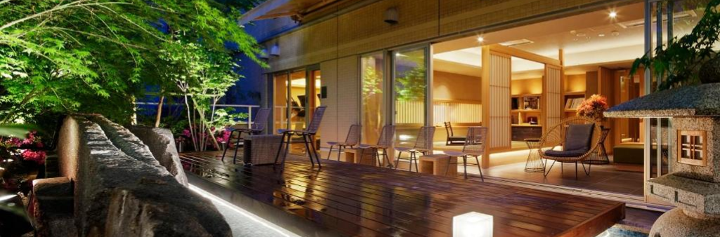 Wooden deck with chairs outside a warmly lit modern house, surrounded by greenery at night.